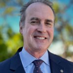 A middle-aged man in a dark suit, blue shirt, and patterned tie smiles outdoors with green trees and blue sky softly blurred in the background. Sourcing, Procurement, 3rd Party Risk