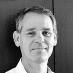 A middle-aged man with short, gray hair is smiling slightly while looking at the camera. He is wearing a light-colored collared shirt and stands in front of a dark, plain background. The black and white image highlights his subtle expression. Sourcing, Procurement, 3rd Party Risk