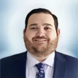 A man with dark hair and a beard, wearing a navy suit, white shirt, and patterned tie, smiles against a light blue, blurred background. Sourcing, Procurement, 3rd Party Risk