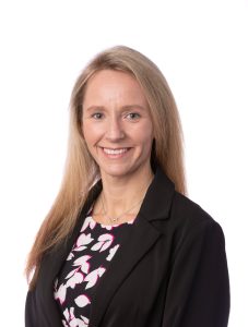 A woman with long blonde hair, wearing a black blazer over a patterned blouse, smiles at the camera against a plain white background. Sourcing, Procurement, 3rd Party Risk