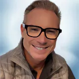 Smiling man with short, light hair wearing black glasses, a grey quilted vest, and a dark shirt, pictured against a soft blue background. Sourcing, Procurement, 3rd Party Risk