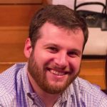 A man with short brown hair and a beard, wearing a light blue checkered shirt, smiles while seated indoors. A blurry handbag and wooden steps are visible in the background. Sourcing, Procurement, 3rd Party Risk