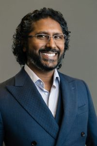 A man with curly dark hair, a beard, and glasses smiles while wearing a blue suit jacket and light-colored shirt, standing in front of a plain grey background. Sourcing, Procurement, 3rd Party Risk
