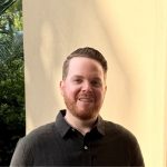 A man with short brown hair and a trimmed beard, wearing a dark button-up shirt, stands smiling in front of a light-colored wall with greenery visible in the background. Sourcing, Procurement, 3rd Party Risk