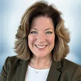 A woman with shoulder-length, wavy light brown hair is smiling at the camera. She is wearing a dark blazer over a white top, and the background is softly blurred in blue and white tones. Sourcing, Procurement, 3rd Party Risk