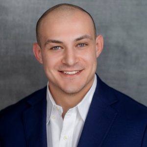 A smiling man with a shaved head, wearing a dark blue suit jacket over a white dress shirt, poses in front of a gray textured background. Sourcing, Procurement, 3rd Party Risk