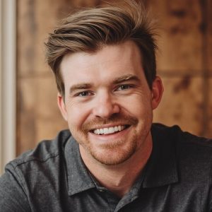 A man with light brown hair and a beard smiles at the camera. He is wearing a dark collared shirt and is in front of a wooden background. Sourcing, Procurement, 3rd Party Risk