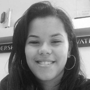 Black and white photo of a young woman with long dark hair, smiling at the camera. She is wearing hoop earrings and a jacket. There are indistinct objects and a partially visible sign in the background. Sourcing, Procurement, 3rd Party Risk
