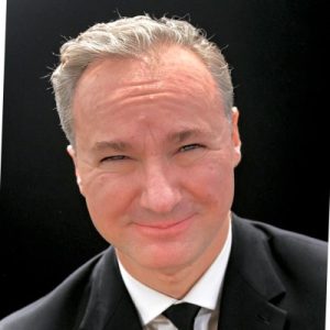 A man with short gray hair, wearing a black suit and tie, smiles at the camera against a plain black background. Sourcing, Procurement, 3rd Party Risk