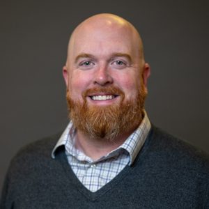 A smiling bald man with a reddish beard, wearing a gray sweater over a checkered shirt, poses in front of a plain dark background. Sourcing, Procurement, 3rd Party Risk