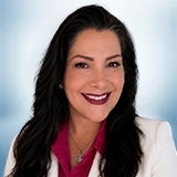 Smiling woman with long dark hair wearing a white blazer over a burgundy blouse, posing in front of a light blue blurred background. Sourcing, Procurement, 3rd Party Risk