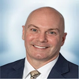 Smiling bald man wearing a dark suit jacket, white shirt, and patterned tie, with a blurred light blue background. Sourcing, Procurement, 3rd Party Risk