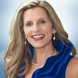 Woman with long, wavy blonde hair, wearing a sleeveless royal blue dress with a ruffled collar, drop earrings, and a necklace, smiling against a blurred background. Sourcing, Procurement, 3rd Party Risk