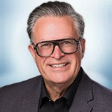 Smiling older man with gray hair and large dark glasses, wearing a dark suit jacket and black shirt, against a light blue background. Sourcing, Procurement, 3rd Party Risk