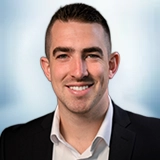 Smiling man with short dark hair wearing a black suit jacket and white shirt, posed in front of a softly blurred blue and white background. Sourcing, Procurement, 3rd Party Risk