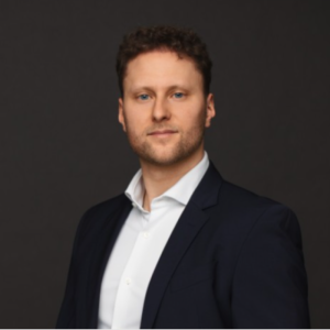 A man with short curly hair and a trimmed beard wears a dark suit jacket and white dress shirt, posing confidently against a plain dark background. Sourcing, Procurement, 3rd Party Risk