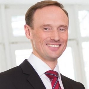 A man in a dark suit, white shirt, and red striped tie smiling in a bright room with large windows in the background. Sourcing, Procurement, 3rd Party Risk