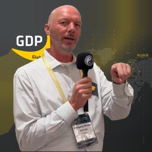 A man in a white shirt holding a microphone stands in front of a backdrop with the word GDP, a world map, and a highlighted Austria. He wears a badge that says Wolfgang Winhofer. Sourcing, Procurement, 3rd Party Risk