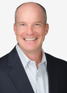 Smiling man with a bald head wearing a dark blazer and a light-colored checkered shirt, posing in front of a white background. Sourcing, Procurement, 3rd Party Risk
