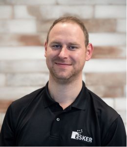 A man with short light brown hair wearing a black Esker polo shirt smiles in front of a blurred light brick wall background. Sourcing, Procurement, 3rd Party Risk