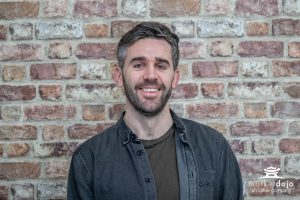 A man with short brown hair and a beard smiles at the camera, standing in front of a brick wall. He is wearing a dark shirt over a t-shirt. A marketdojo logo appears in the lower right corner. Sourcing, Procurement, 3rd Party Risk