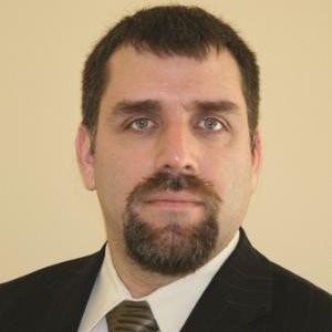 A man with short dark hair, a beard, and mustache, wearing a black pinstripe suit, white shirt, and striped tie, poses in front of a plain beige background. Sourcing, Procurement, 3rd Party Risk