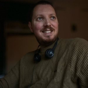 A man with short hair and a beard, wearing a brown sweater and black headphones around his neck, is smiling and looking to the side in a dimly lit indoor setting. Sourcing, Procurement, 3rd Party Risk