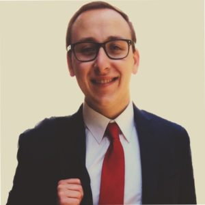 Smiling person wearing glasses, a dark suit, white shirt, and red tie, holding a jacket over their shoulder, posed against a plain light background. Sourcing, Procurement, 3rd Party Risk