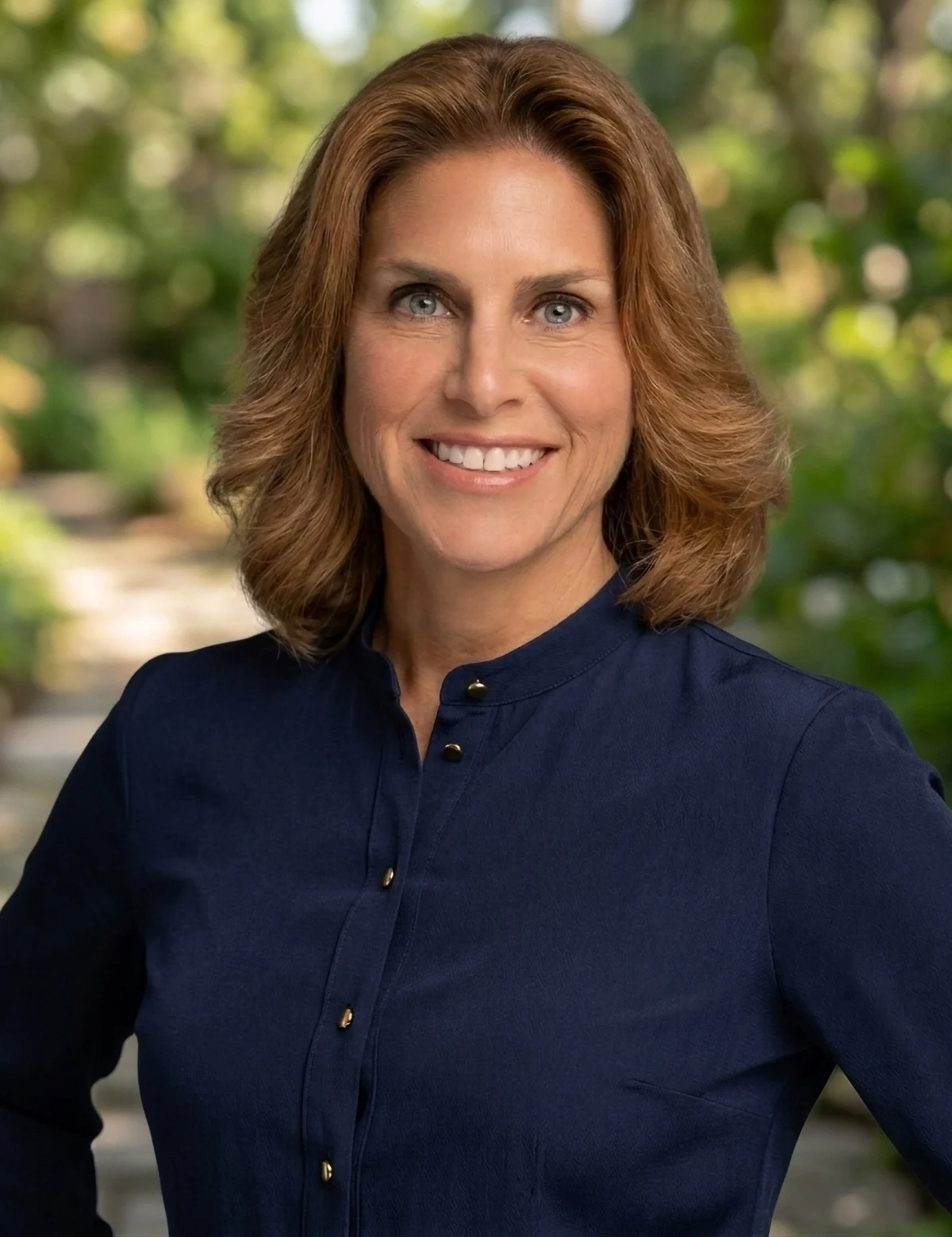 A woman with shoulder-length brown hair wearing a navy button-up shirt smiles outdoors, with a blurred background of greenery and sunlight. Sourcing, Procurement, 3rd Party Risk