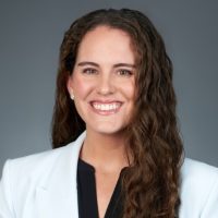 A woman with long, wavy brown hair smiles at the camera. She is wearing a white blazer over a black top and small stud earrings, standing against a simple gray gradient background. Sourcing, Procurement, 3rd Party Risk